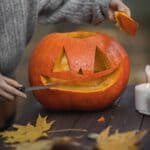 The 5 Best Pumpkin Carving Kits for Tippah County Families A person carving a pumpkin into a jack-o'-lantern on an outdoor wooden table with autumn leaves.